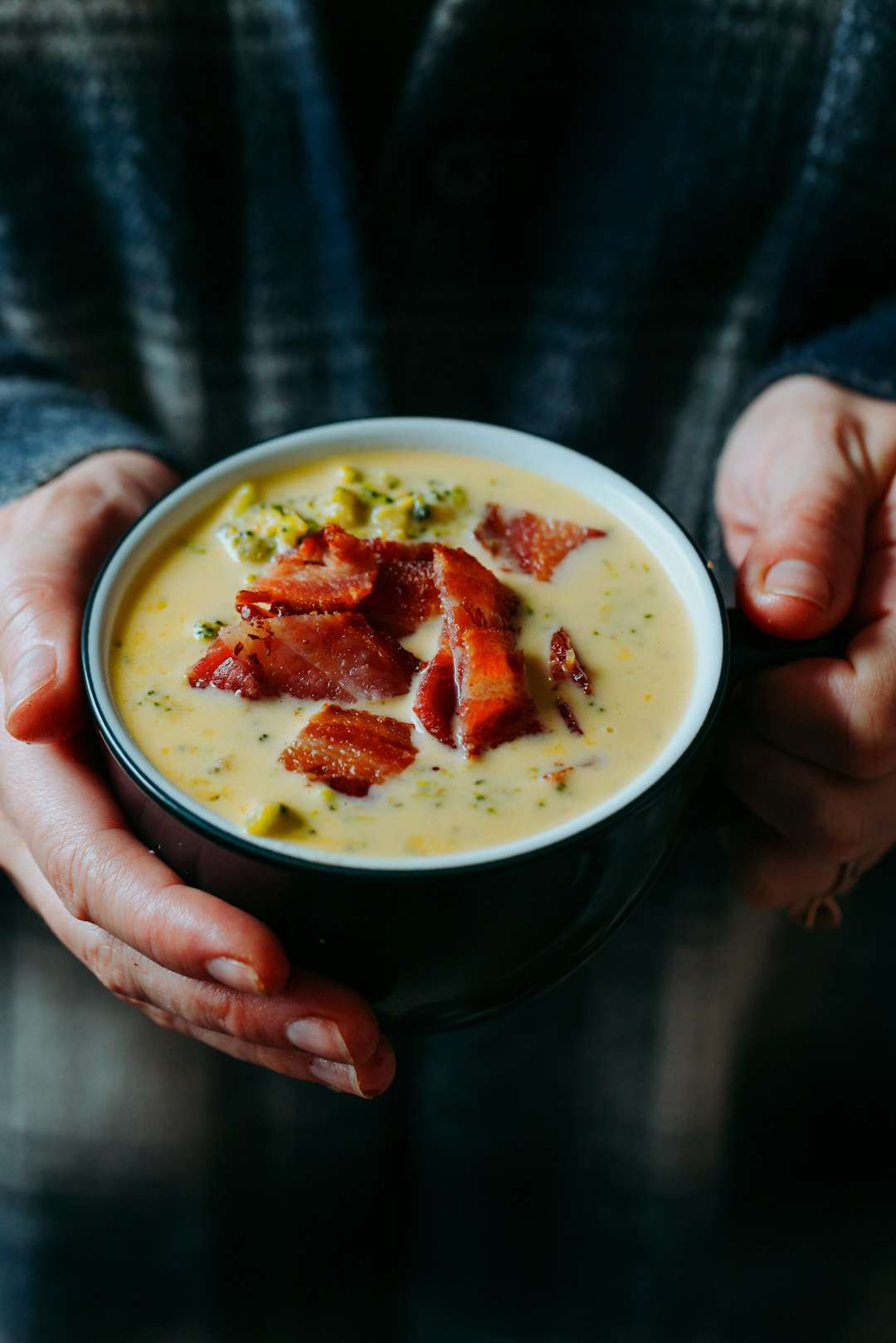 A person holding a cozy bowl of creamy corn chowder topped with crispy bacon bits
